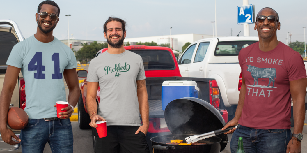 Three men standing around a table with food and drinks in a parking lot.